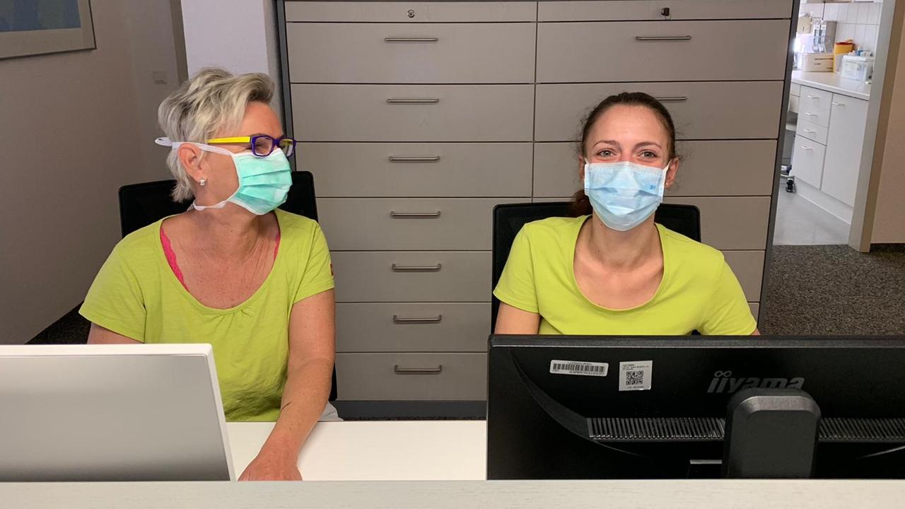 Eine Sprechstundenhilfe und eine Arzthelferin der Praxis Heinert im badischen Bahlingen sitzen hinter dem Anmeldungstresen. Sie tragen Mundschutzmasken. Eine Sprechstundenhilfe und eine Arzthelferin der Praxis Heinert im badischen Bahlingen sitzen hinter dem Anmeldungstresen. Sie tragen Mundschutzmasken.