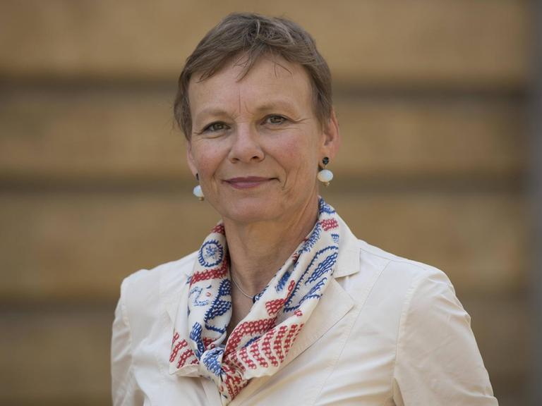 Sabine Kunst, Frau mit kurzen Haaren steht in heller Jacke vor einem hellbraunen Hintergrund und lächelt in die Kamera.
