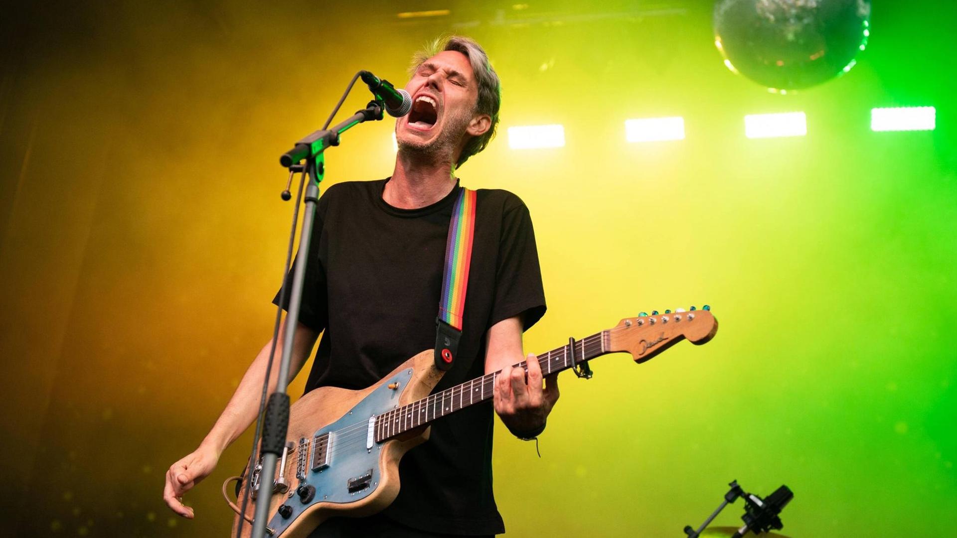 Dirk von Lowtzow steht mit E-Gitarre und singt in ein Standmikrofon, im Hintergrund ist ein gelb-grüner Farblichtverlauf und eine Diskokugel.