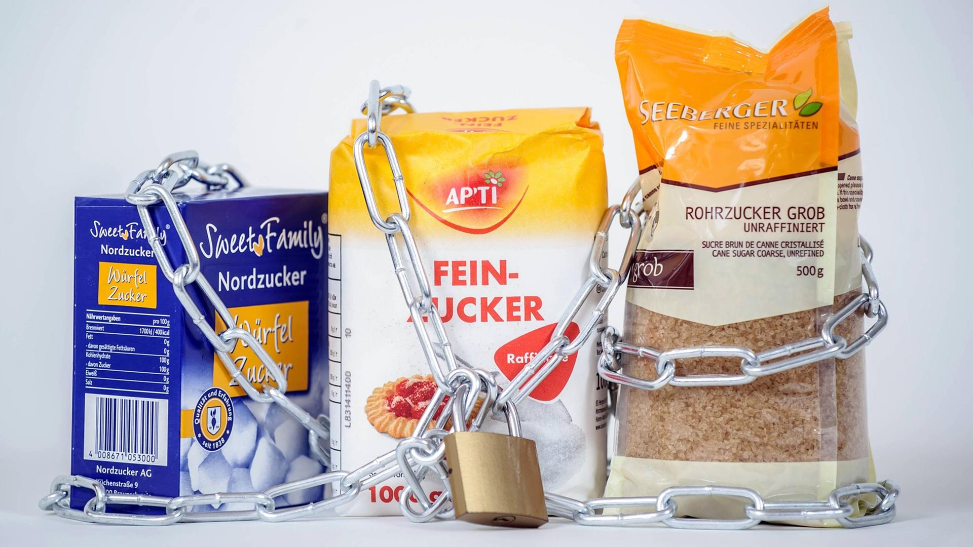 Eine Packung weißer Zucker (M), Würfelzucker (l) und brauner Rohzucker werden von einer Eisenkette samt Schloss zusammen gehalten. Eine Packung weißer Zucker (M), Würfelzucker (l) und brauner Rohzucker werden von einer Eisenkette samt Schloss zusammen gehalten.