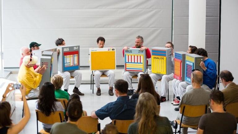 Eine Performance von Künstlern, die mit bunten Pappkartons im Halbkreis vor Zuschauern sitzen Eine Performance von Künstlern, die mit bunten Pappkartons im Halbkreis vor Zuschauern sitzen