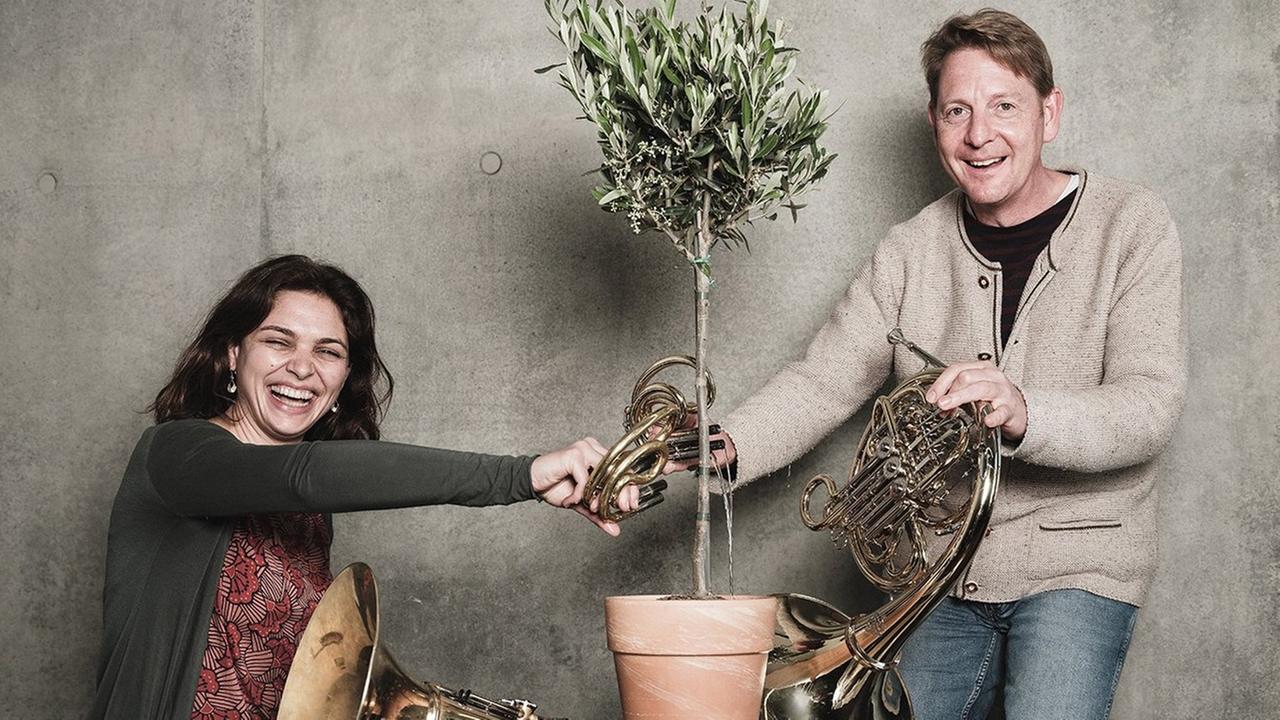 Eine Frau und ein Mann lachen in die Kamera. Zwischen ihnen steht ein Olivenbaum, den sie mit einem kleinen Horn gießen. Beide haben jeweils ein großes Horn in der Hand. Eine Frau und ein Mann lachen in die Kamera. Zwischen ihnen steht ein Olivenbaum, den sie mit einem kleinen Horn gießen. Beide haben jeweils ein großes Horn in der Hand.