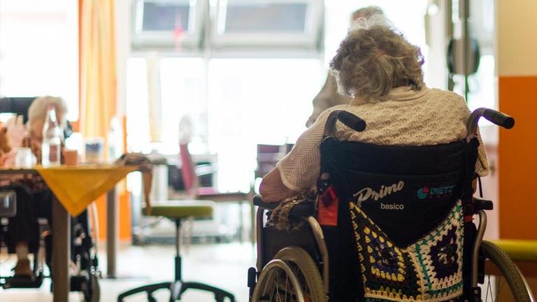 Eine Frau im Rollstuhl sitzt allein im Gemeinschaftsraum eines Altenheims.