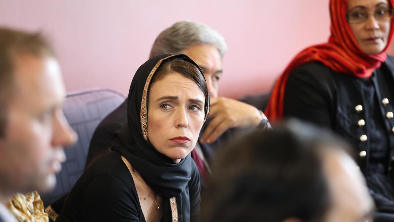 Jacinda Adern schaut ernst, hat sich ein schwarzes Tuch um den Kopf geschlungen und sitzt mit anderen Personen in einem Raum. Jacinda Adern schaut ernst, hat sich ein schwarzes Tuch um den Kopf geschlungen und sitzt mit anderen Personen in einem Raum.