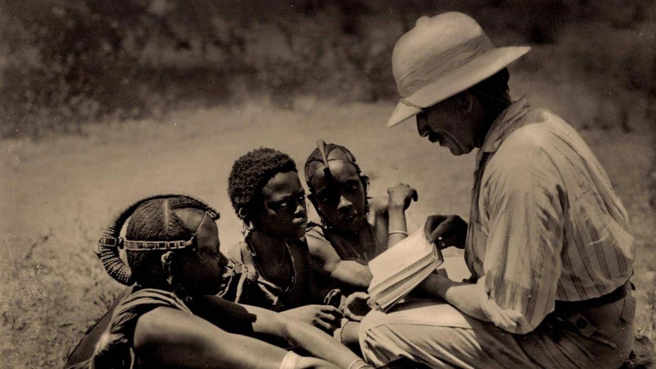 Eine historische Aufnahme von 1935 zeigt einen Missionar, der drei knieenden Massai aus erhöhter Perspektive das Neue Testament zeigt.