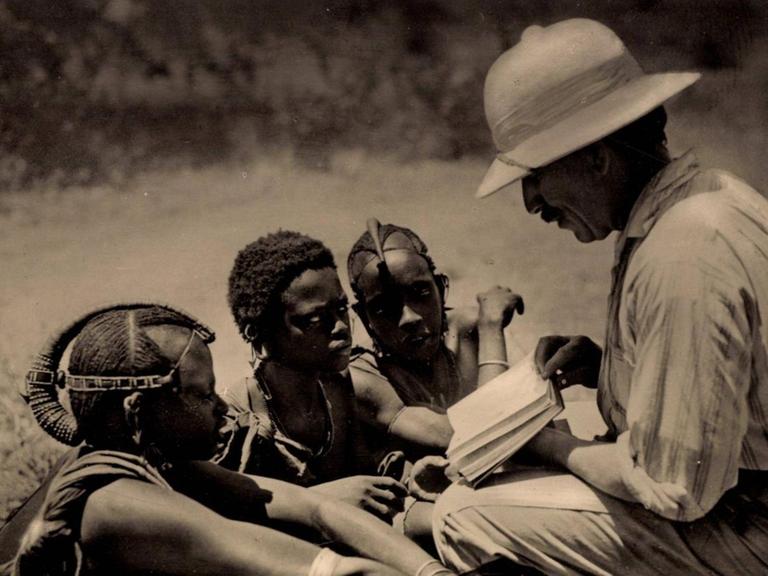 Eine historische Aufnahme von 1935 zeigt einen Missionar, der drei knieenden Massai aus erhöhter Perspektive das Neue Testament zeigt.