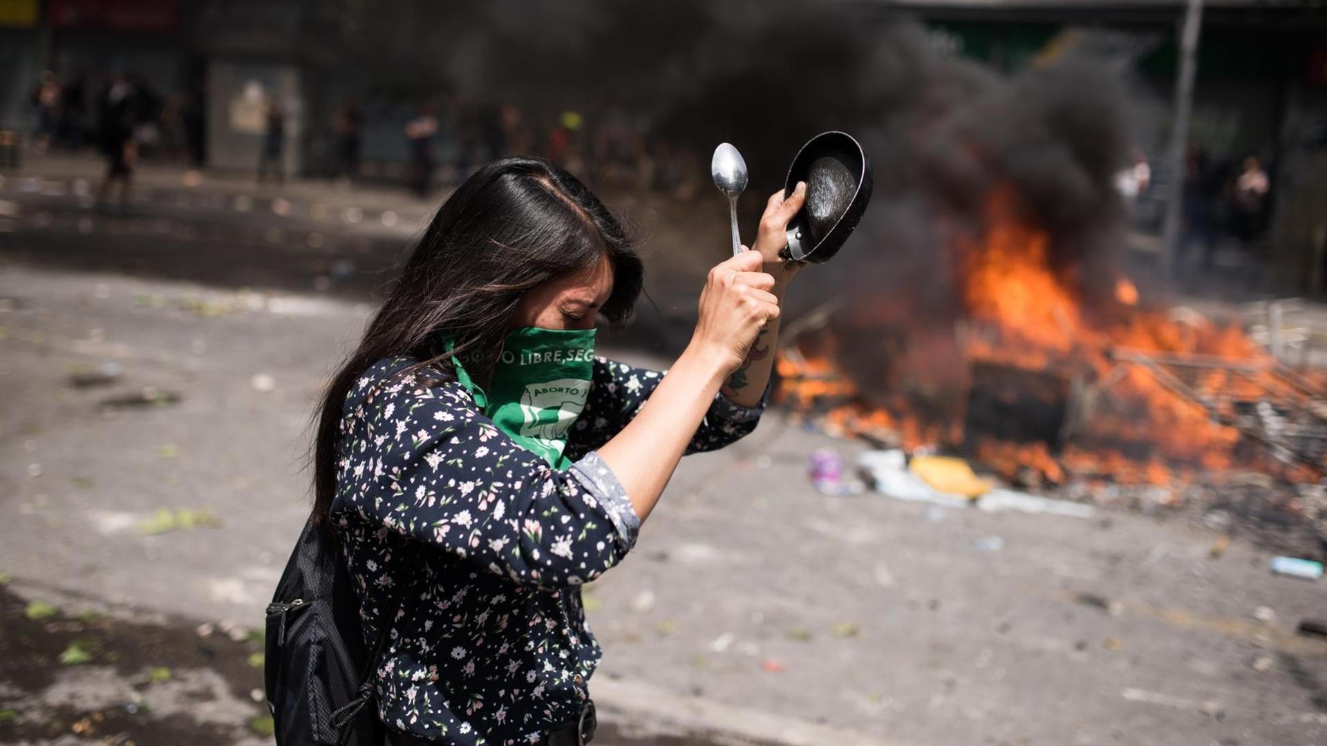 Proteste in Chile gegen Preiserhöhungen im öffentlichen Nahverkehr. Eine Frau schlägt auf ein Gefäß, im Hintergrund brennender Müll auf der Straße.