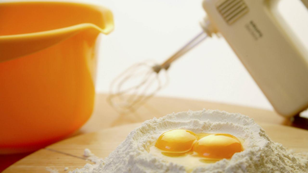 Zwei Eigelb auf Mehl sind vor einer Schüssel und einem Mixer zu sehen. Zwei Eigelb auf Mehl sind vor einer Schüssel und einem Mixer zu sehen.