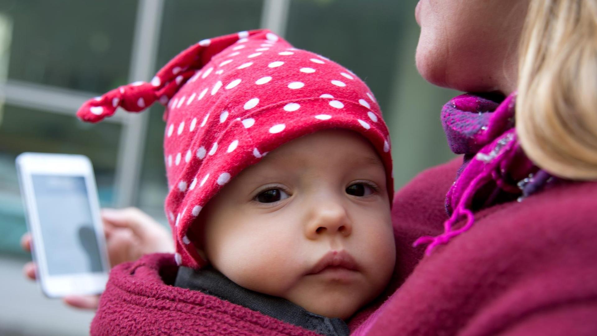Eine Frau schaut auf ihr Smartphone, während sie ihre Tochter in einer Babytrage vor dem Bauch trägt. Eine Frau schaut auf ihr Smartphone, während sie ihre Tochter in einer Babytrage vor dem Bauch trägt.