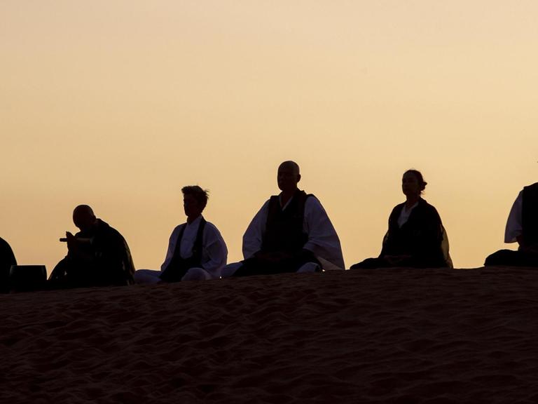 Silhouetten von sitzenden Menschen, die während der Dämmerung auf einer Düne meditieren.