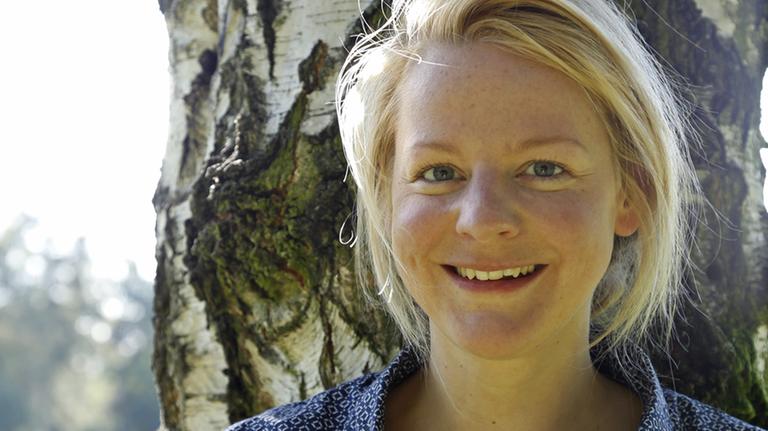 Anna Leuschner lehnt an einer Birke und lächelt in die Kamera