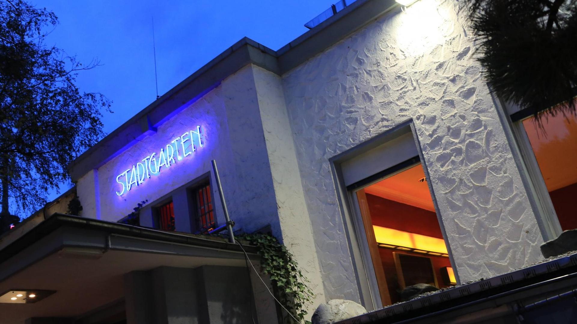 Ansicht einer angeleuchteten, weißen Hausfassade in der Dämmerung mit einem überdachten Eingang. Über dem Eingang leuchtet im blauen Neonlicht der Name "Stadtgarten". Ansicht einer angeleuchteten, weißen Hausfassade in der Dämmerung mit einem überdachten Eingang. Über dem Eingang leuchtet im blauen Neonlicht der Name "Stadtgarten".