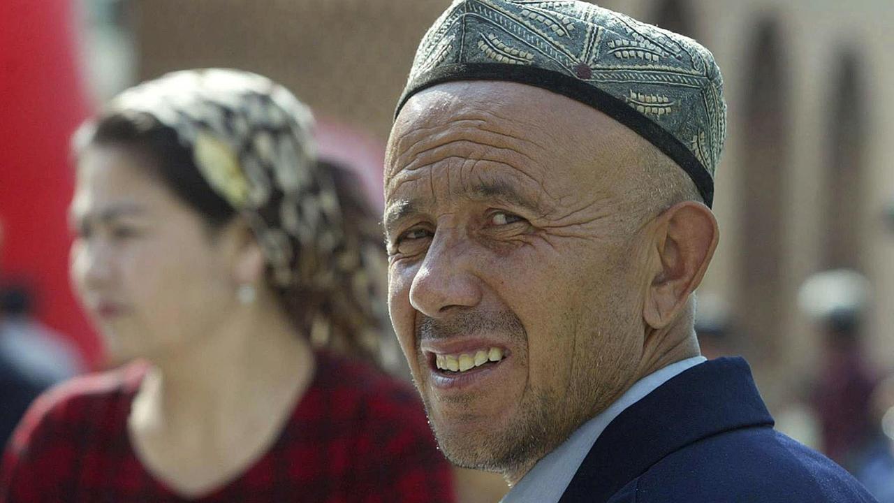 Ein uigurischer Mann mit traditioneller Kopfbedeckung und verschwommen im Hintergrund eine Frau in traditioneller Kleidung. Ein uigurischer Mann mit traditioneller Kopfbedeckung und verschwommen im Hintergrund eine Frau in traditioneller Kleidung.