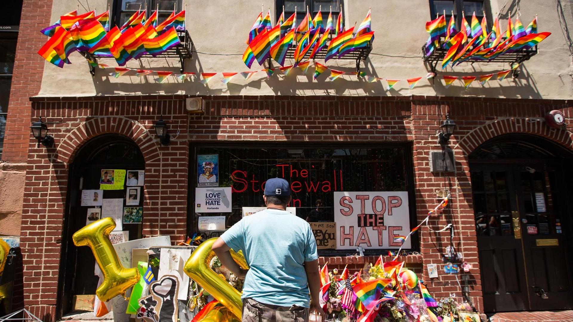 Ein Mann steht vor dem Stonewall Inn. Vor dem Gebäude liegen viele Gedenkobjekte. An der Fassade hängen Flaggen. Ein Mann steht vor dem Stonewall Inn. Vor dem Gebäude liegen viele Gedenkobjekte. An der Fassade hängen Flaggen.