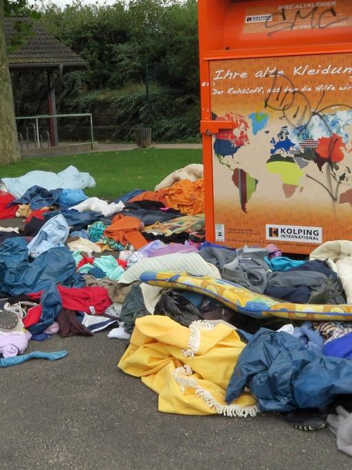 Textilien, Schuhe und Polster liegen neben einem Container für Altkleider auf der Straße.