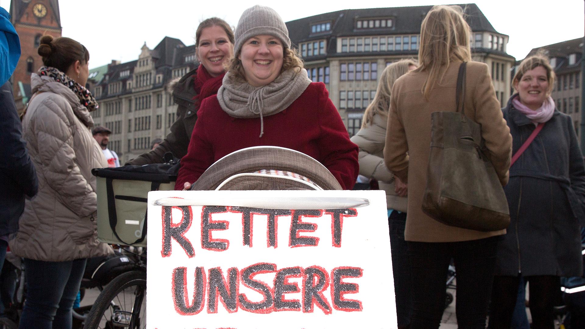 Eine Frau hat ein Schild an ihrem Kinderwagen mit der Aufschrift "Rettet unsere Hebammen" befestigt. Eine Frau hat ein Schild an ihrem Kinderwagen mit der Aufschrift "Rettet unsere Hebammen" befestigt.