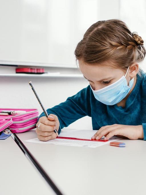 Die achtjährige Anna sitzt mit Maske an einem Tisch und bearbeitet ein Übungsblatt.