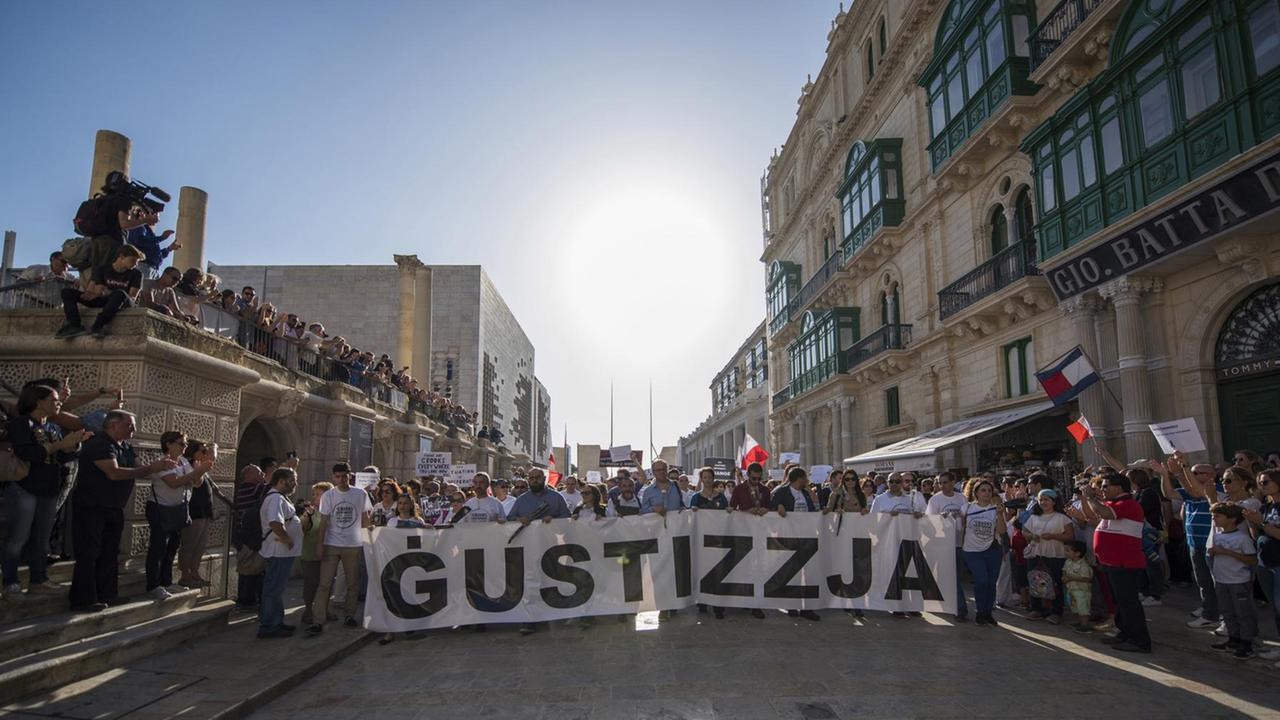 Die Menschen stehen quer über eine gepflasterte Straße hinter einem breiten Transparent mit der Aufschrift "Gustizzja". Auch an den Seiten stehen Demonstranten sowie Medienvertreter. Die Menschen stehen quer über eine gepflasterte Straße hinter einem breiten Transparent mit der Aufschrift "Gustizzja". Auch an den Seiten stehen Demonstranten sowie Medienvertreter.