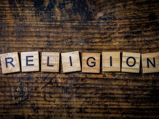 Das Wort "Religion" ist aus hellen Buchstaben-Holzplättchen auf ein dunkles Holzbrett gelegt.