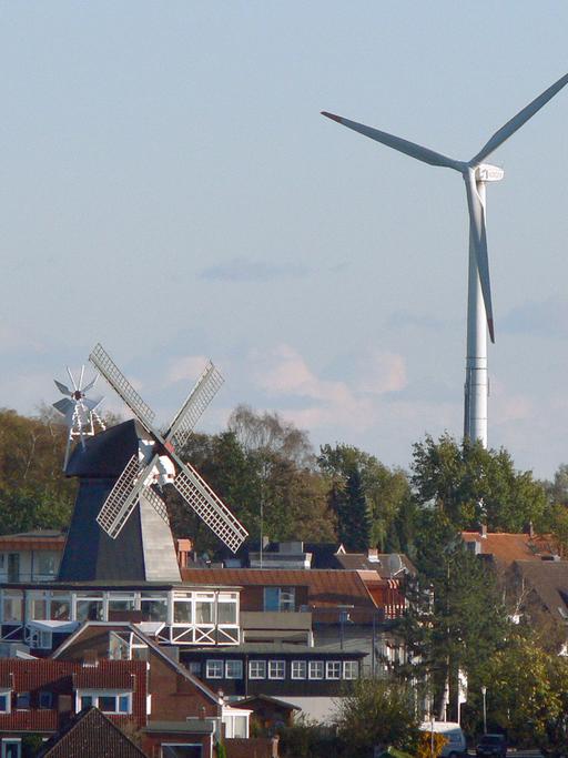 Eine alte Windmühle steht zwischen Wohnhäusern in Kiel, im Hintergrund ein modernes Windrad.