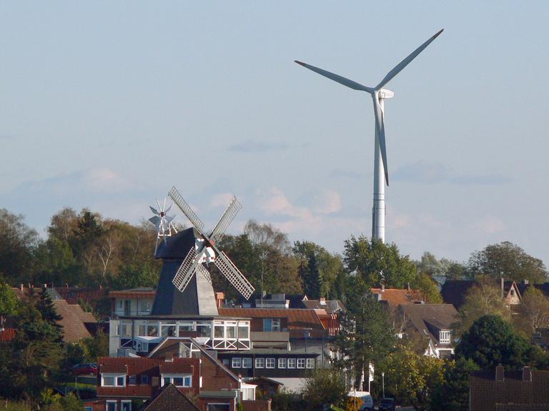 Eine alte Windmühle steht zwischen Wohnhäusern in Kiel, im Hintergrund ein modernes Windrad.