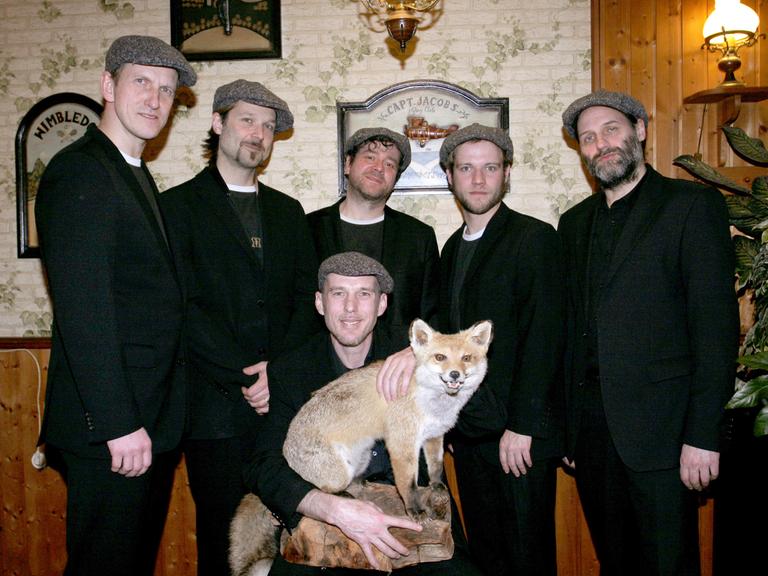 Die Mitglieder der Band "Jazzkantine" stehen zusammen in schwarzen Anzügen. Einer kniet in der Mitte und hält einen ausgestopften Fuchs in den Händen.