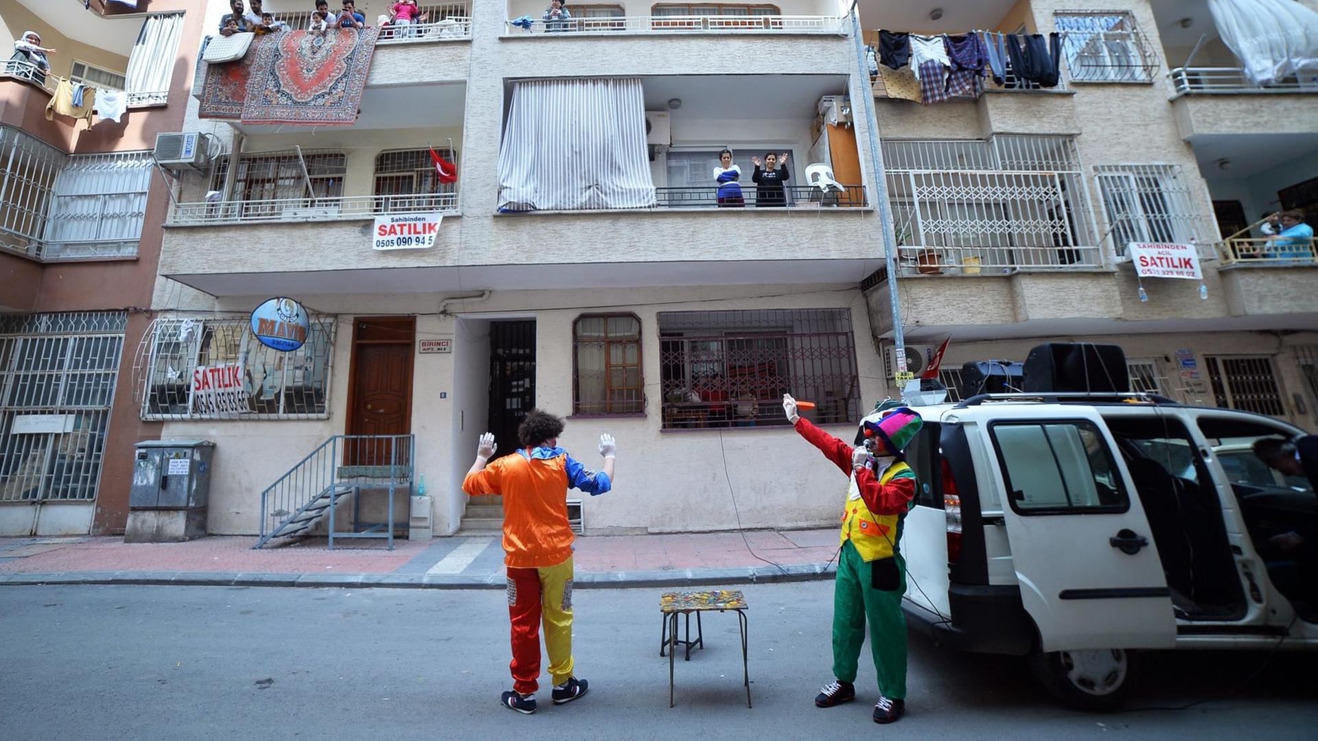Zwei Clowns stehen neben einem Bulli in einer Wohnstraße, auf den Balkons lachen Kinder und hängt Wäsche. Zwei Clowns stehen neben einem Bulli in einer Wohnstraße, auf den Balkons lachen Kinder und hängt Wäsche.