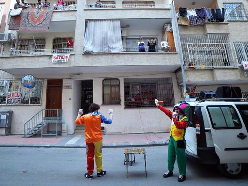 Zwei Clowns stehen neben einem Bulli in einer Wohnstraße, auf den Balkons lachen Kinder und hängt Wäsche.