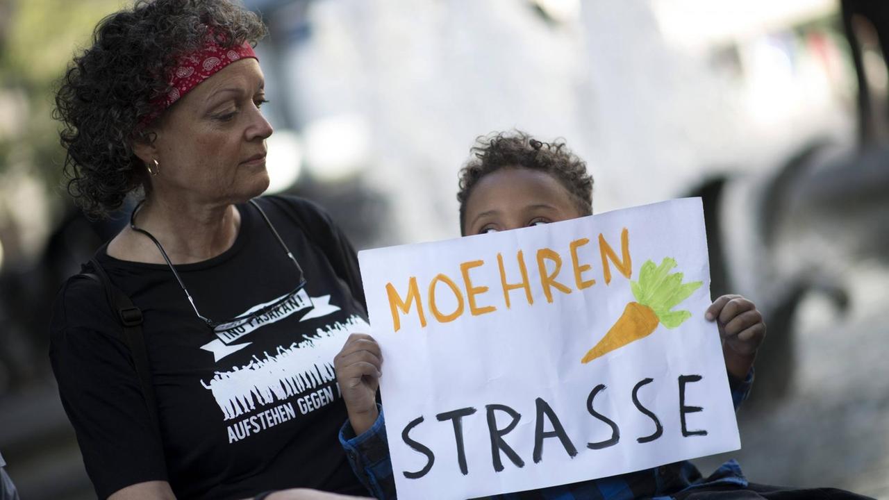 Ein Kind hält ein Schild mit einer gemalten Möhre über dem Text "Moehrenstraße" hoch, anlässlich des Umbenennungsfestes von Mohrenstraße zu Anton W Amo Strasse Ein Kind hält ein Schild mit einer gemalten Möhre über dem Text "Moehrenstraße" hoch, anlässlich des Umbenennungsfestes von Mohrenstraße zu Anton W Amo Strasse