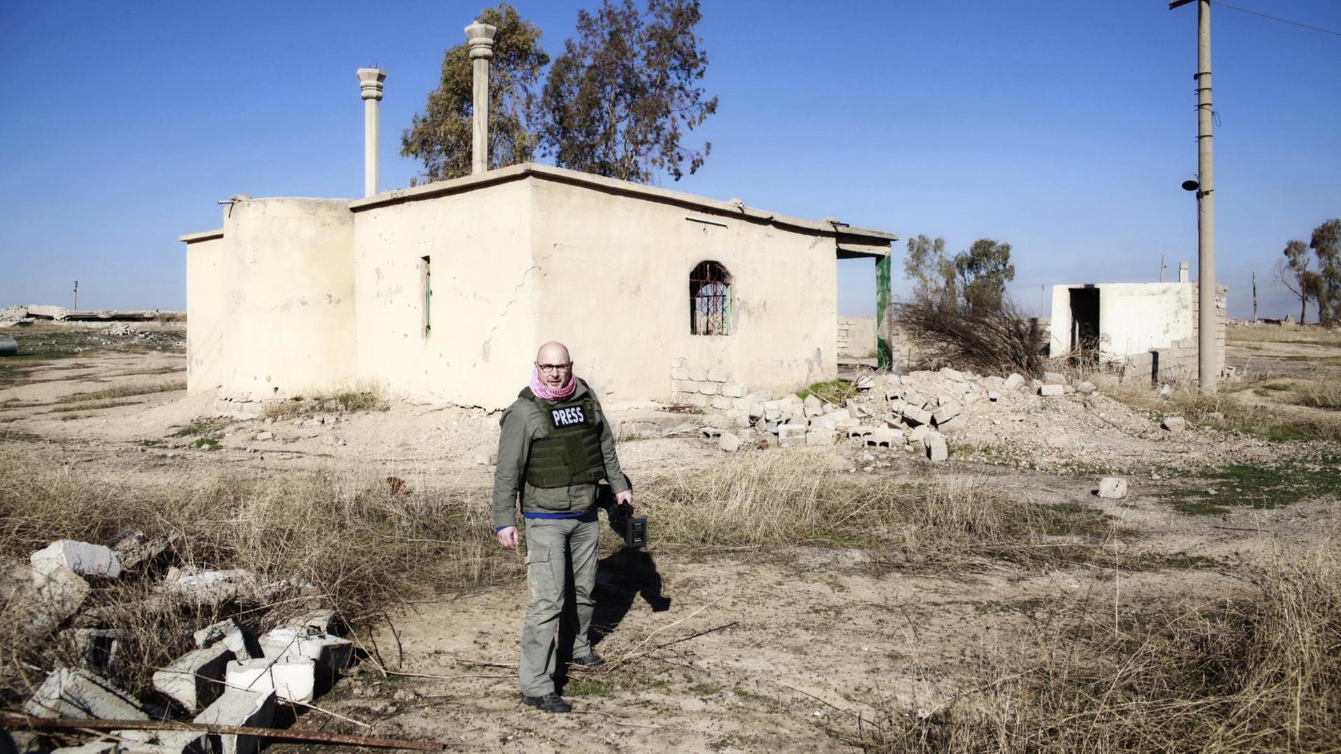 Ein glatzköpfiger Mann mit Brille steht vor einem teilweise zerstörten weißen Gebäude in einer Landschaft voller Geröll und verdorrtem Gras. In der Hand hält er eine Filmkamera. Ein glatzköpfiger Mann mit Brille steht vor einem teilweise zerstörten weißen Gebäude in einer Landschaft voller Geröll und verdorrtem Gras. In der Hand hält er eine Filmkamera.