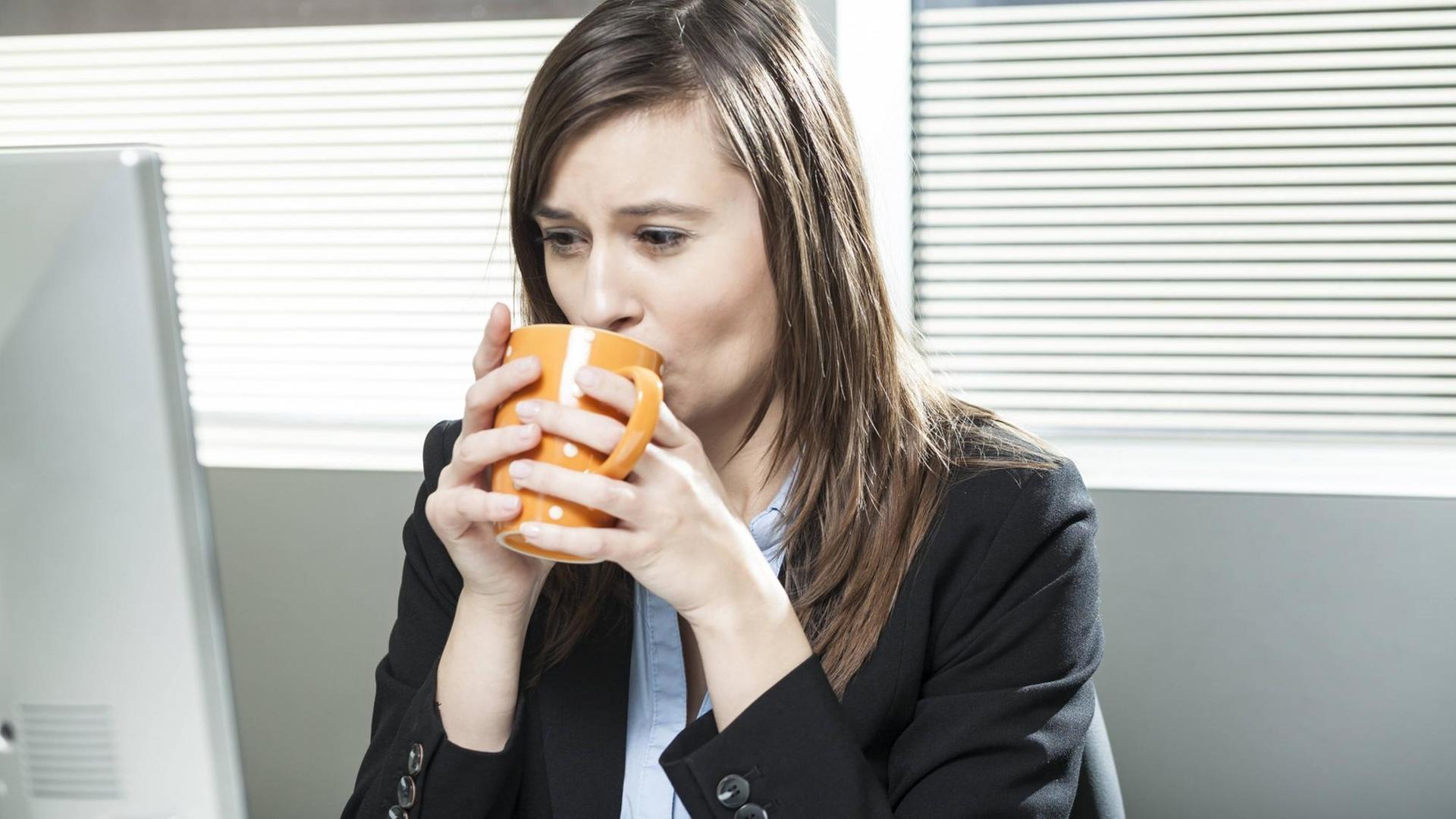 Farbfoto: Frau vor dem Computerbildschirm trinkt eine Tasse Kaffee im Büro
