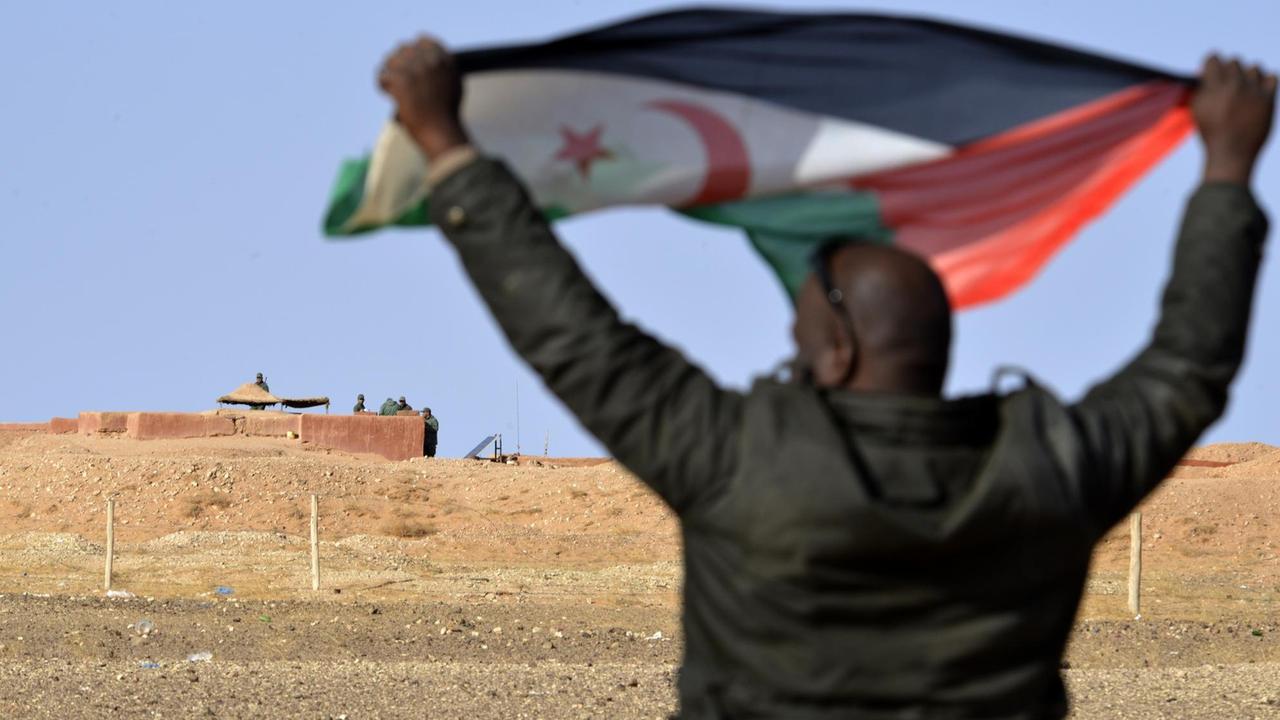 Ein Mann aus dem Volk der Sahrauis hält eine Flagge der Widerstandsorganisation Polisario hoch, im Hintergrund sichern marokkanische Soldaten die Grenzmauer zum von Marokko kontrollierten Teil der Westsahara Ein Mann aus dem Volk der Sahrauis hält eine Flagge der Widerstandsorganisation Polisario hoch, im Hintergrund sichern marokkanische Soldaten die Grenzmauer zum von Marokko kontrollierten Teil der Westsahara