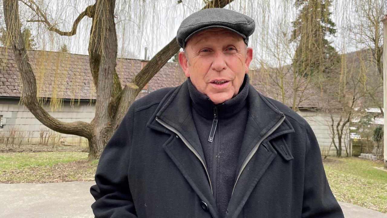 Ein älterer Mann mit Schirmmütze steht im Freien vor einem kleinen Gebäude. Ein älterer Mann mit Schirmmütze steht im Freien vor einem kleinen Gebäude.