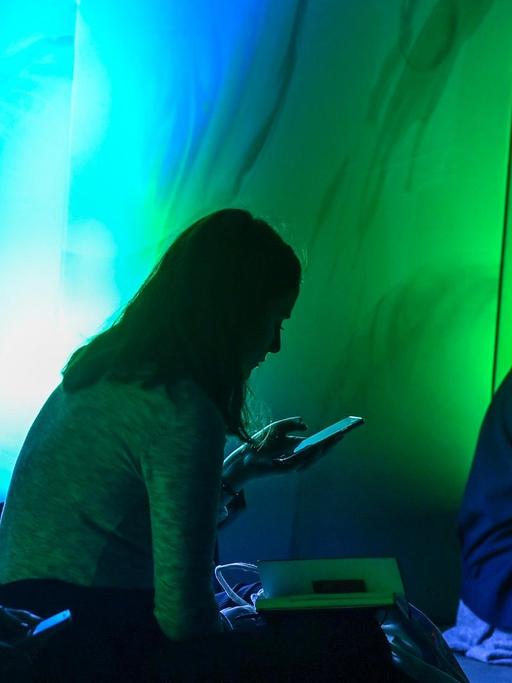 Drei Personen sitzen in einem grünblau dunkel erleuchtetem Raum mit ihren Smartphones in der Hand.