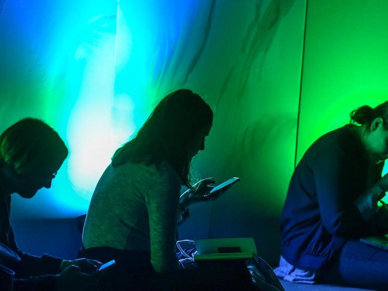 Drei Personen sitzen in einem grünblau dunkel erleuchtetem Raum mit ihren Smartphones in der Hand.