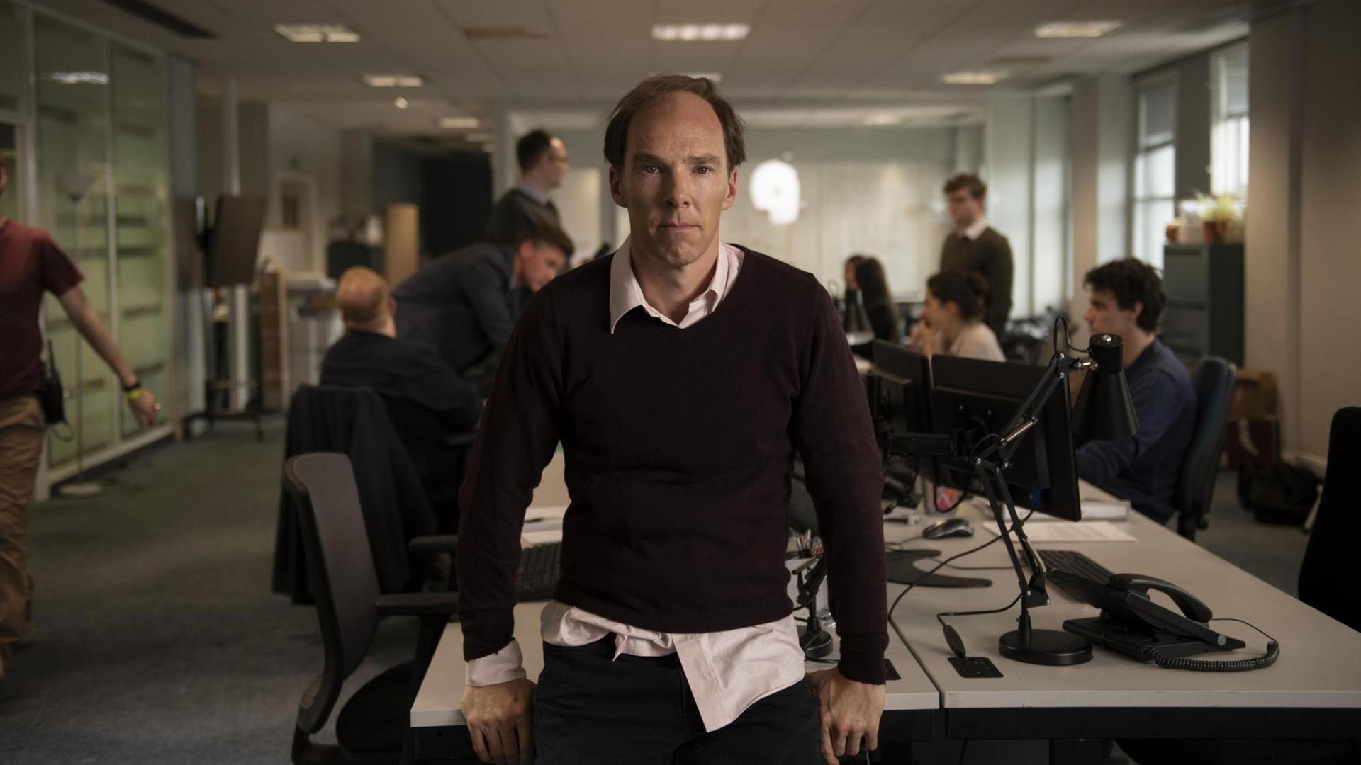 Benedict Cumberbatch in der Rolle des Dominic Cummings in dem Film "Brexit - The uncivil war" sitzt mittig auf einem Tisch in einem geschäftigen Großraumbüro und blickt in die Kamera.