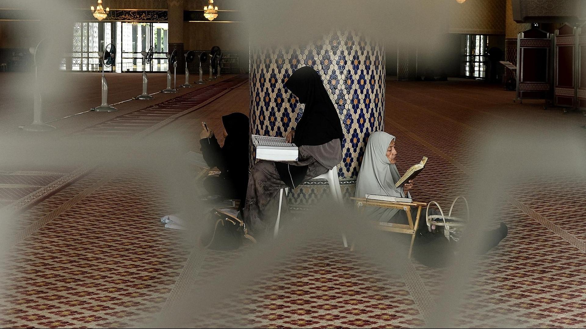 Drei verschleierte Frauen sitzen in einer Moschee gegen eine Säule gelehnt und lesen den Koran.
