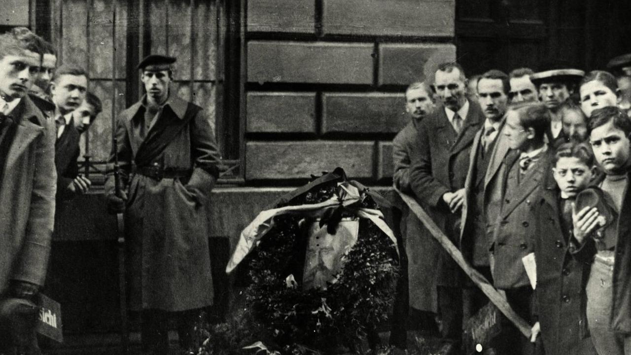 An der Stelle, an der Kurt Eisner in München auf offener Straße ermordet wurde, liegen Blumenkränze. Ringsum stehen Menschen und trauern. An der Stelle, an der Kurt Eisner in München auf offener Straße ermordet wurde, liegen Blumenkränze. Ringsum stehen Menschen und trauern.
