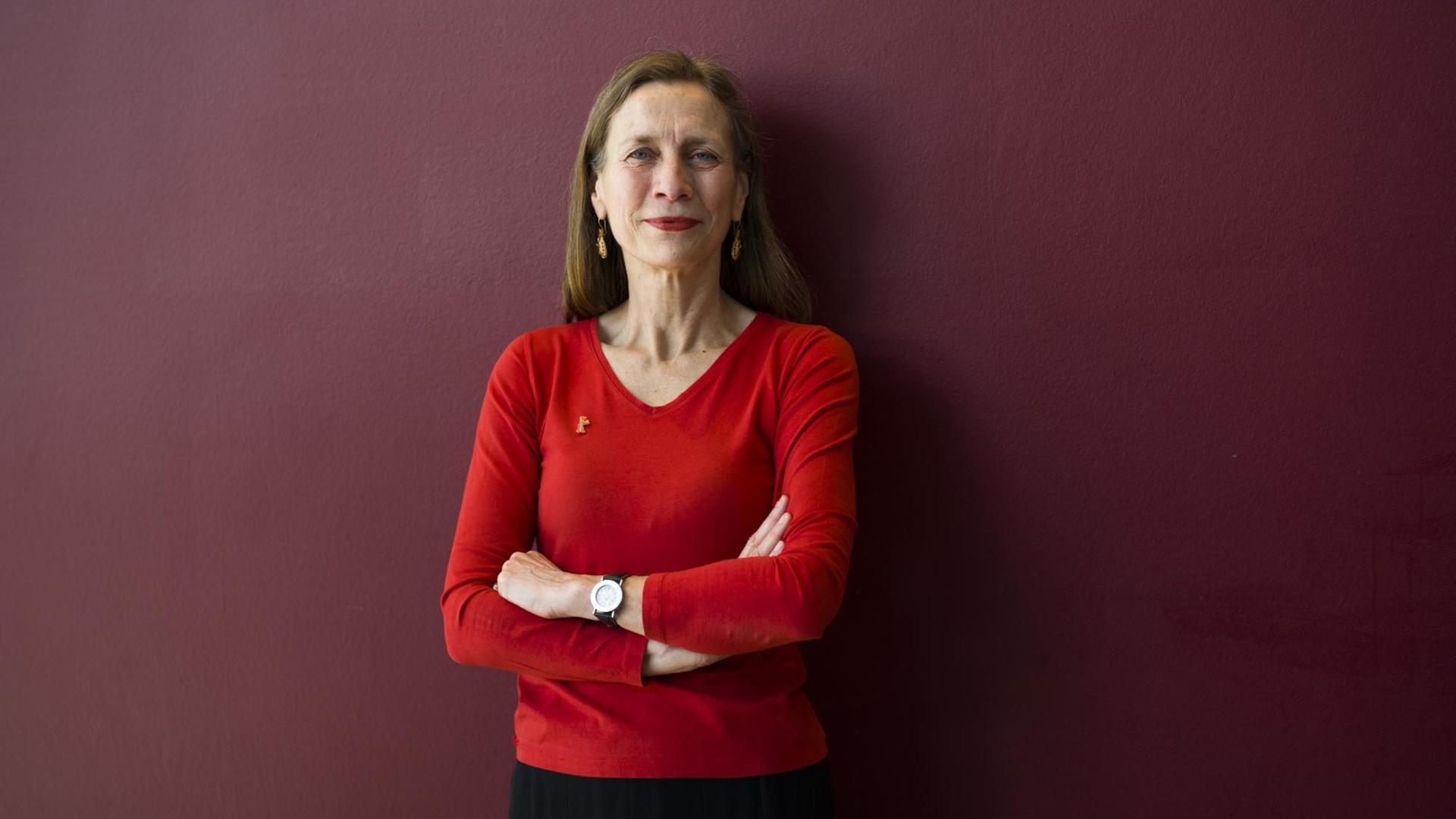 Mariette Rissenbeek steht mit verschränkten Armen in einem roten Oberteil vor einer dunkelroten Wand. Mariette Rissenbeek steht mit verschränkten Armen in einem roten Oberteil vor einer dunkelroten Wand.