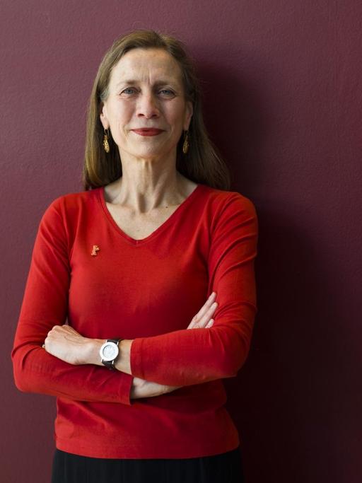 Mariette Rissenbeek steht mit verschränkten Armen in einem roten Oberteil vor einer dunkelroten Wand.