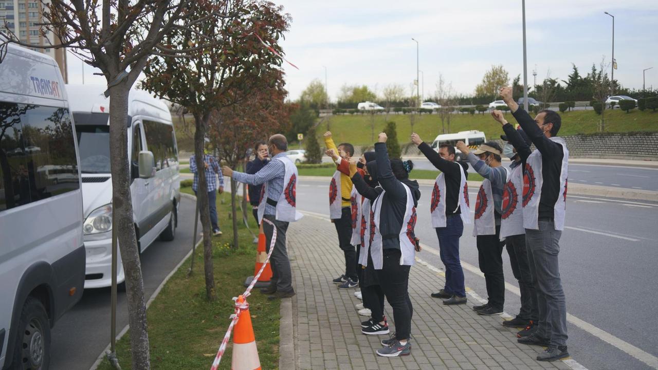 Eine Gruppe Menschen mit weißen ärmellosen Umhängen stehen am Straßenrand und recken die Fäuste gegen vorbeifahrende Busse. Eine Gruppe Menschen mit weißen ärmellosen Umhängen stehen am Straßenrand und recken die Fäuste gegen vorbeifahrende Busse.