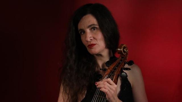 Die Musikerin steht in Abendkleidung und offenem schwarzen, langem Haar mit ihrem Instrument vor rotem Hintergrund und schaut in die Kamera.