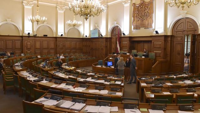 Das lettische Parlament in Riga von innen: Auf den Bänken sitzen einige Abgeordnete, vor dem Rederpult unterhalten sich zwei Frauen und zwei Männer.