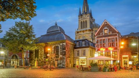 Die Gebäude am Fischmarkt in der Aachener Altstadt liegen hell erleuchtet in der abendlichen Dämmerung, davor sitzen Leute in Restaurants.