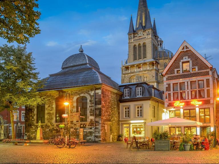 Die Gebäude am Fischmarkt in der Aachener Altstadt liegen hell erleuchtet in der abendlichen Dämmerung, davor sitzen Leute in Restaurants.