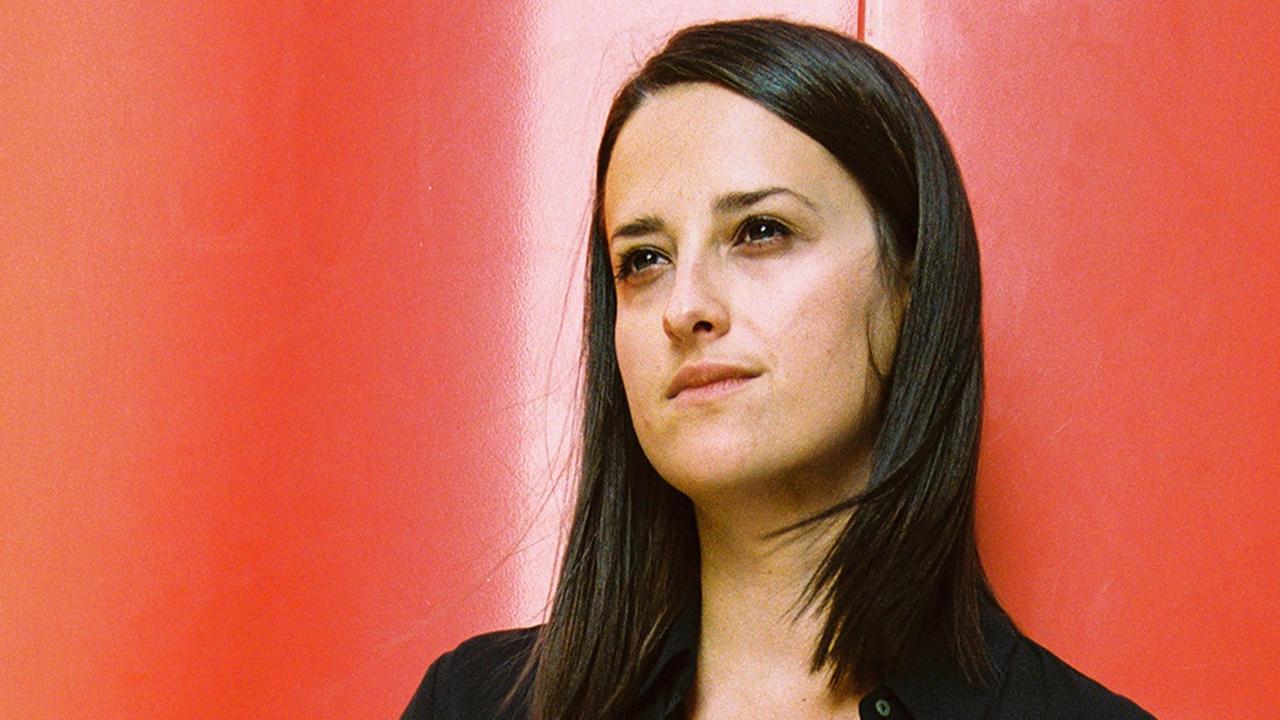Eine Frau mit langen, schwarzen Haaren ist im Porträt vor einer rot-orangenen Wand zu sehen. Sie blickt leicht nach oben und nach rechts aus dem Bild. Eine Frau mit langen, schwarzen Haaren ist im Porträt vor einer rot-orangenen Wand zu sehen. Sie blickt leicht nach oben und nach rechts aus dem Bild.