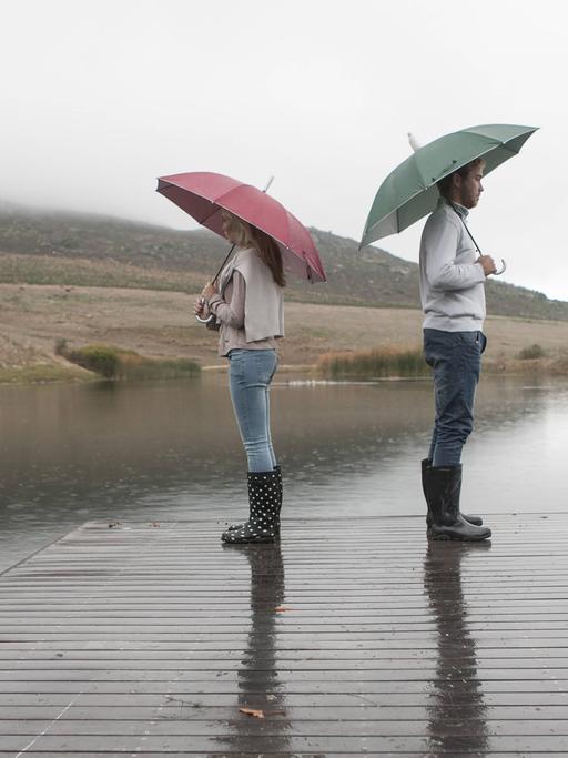 Eine Frau und ein Mann stehen voneinander abgewendet mit Schirmen auf einem hölzernen Steg im Regen.