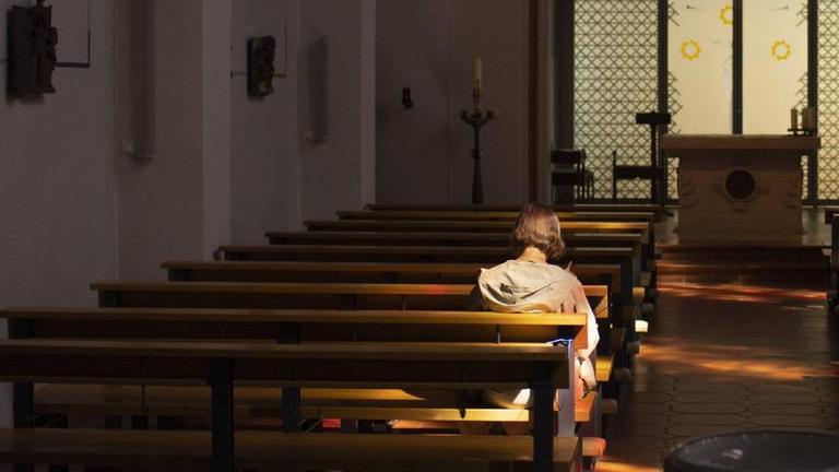 Eine Frau sitzt alleine auf einer Kirchenbank und wird von einem Lichtstrahl, der durch Fenster fällt, angestrahlt. Eine Frau sitzt alleine auf einer Kirchenbank und wird von einem Lichtstrahl, der durch Fenster fällt, angestrahlt.