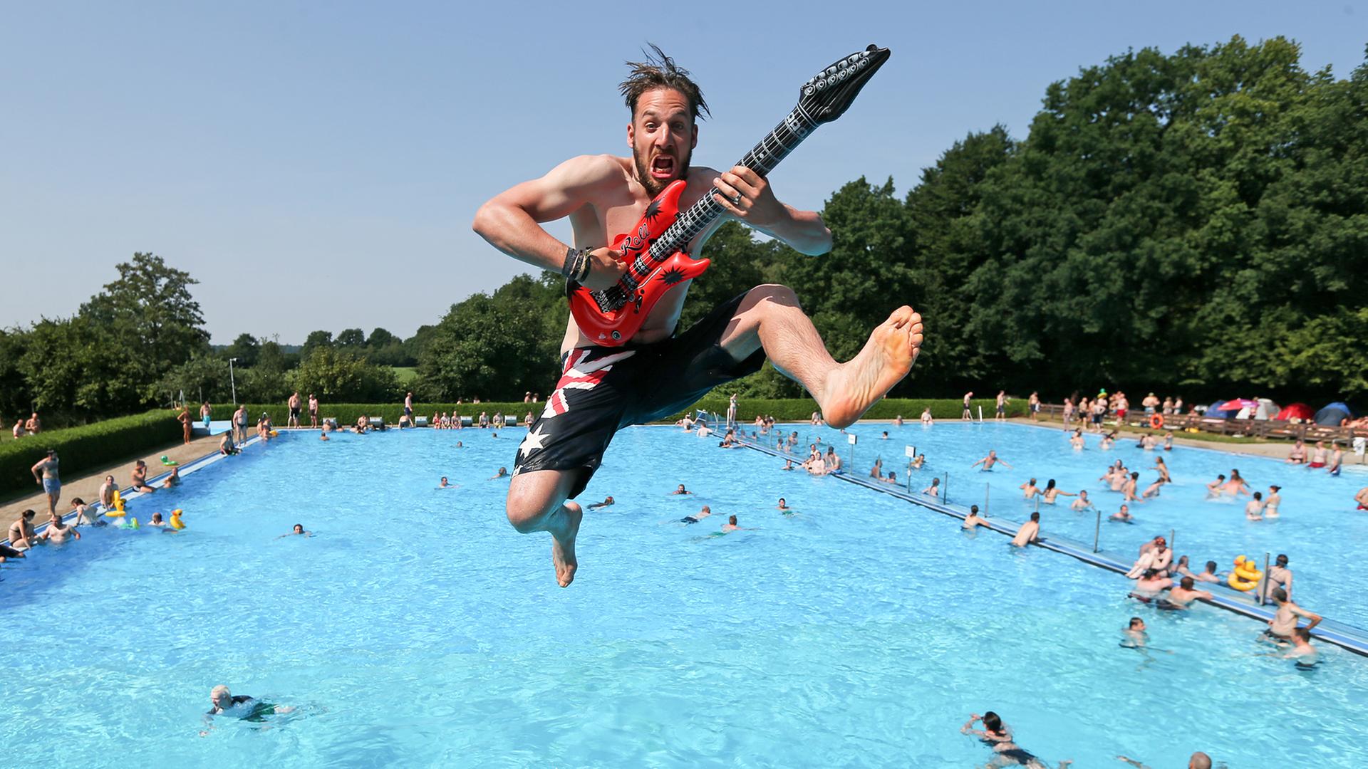 Ein Mann mit einer Plastik-Gitarre in der Hand spring in ein Schwimm-Becken.