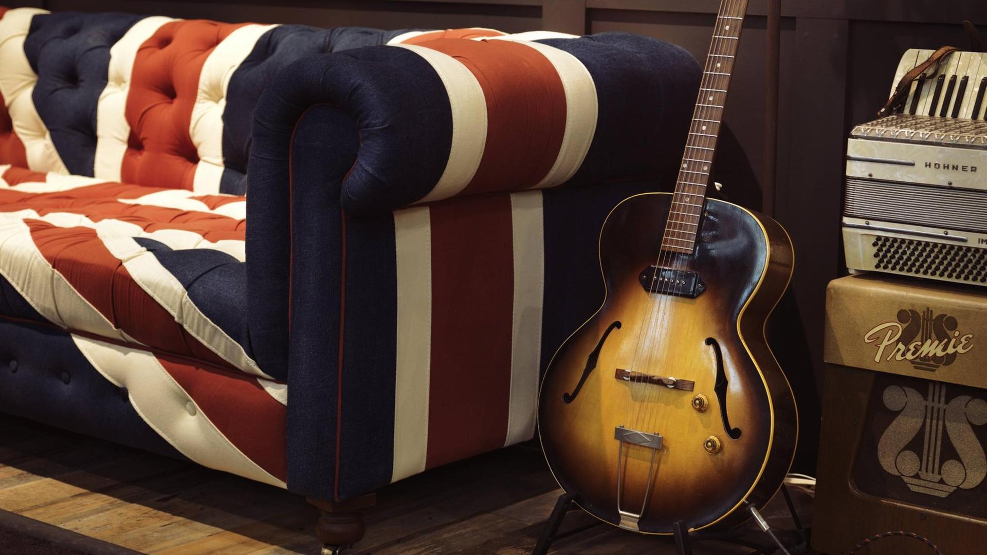 Eine alte akustische Gibson Gitarre steht auf einem Ständer neben einem Sofa mit dem Union Jack als Muster. Eine alte akustische Gibson Gitarre steht auf einem Ständer neben einem Sofa mit dem Union Jack als Muster.